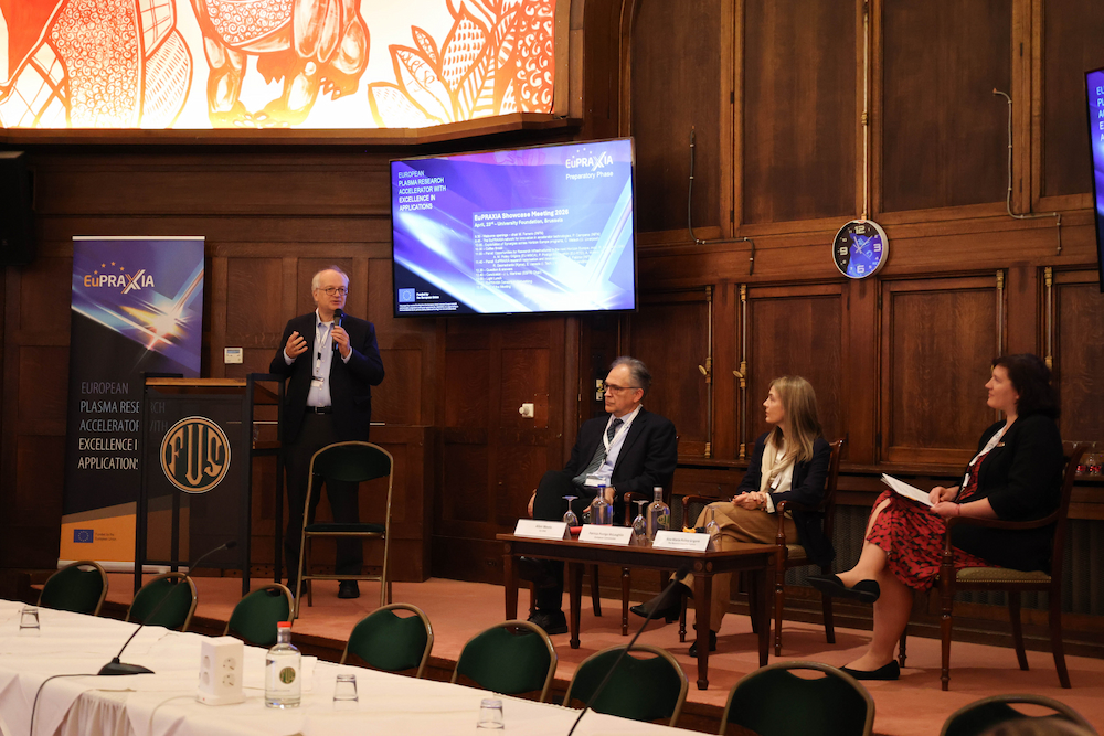 Four people on a stage, with on standing and the other three sitting on chairs. A large screen is mounted to the wall.