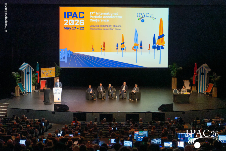Five panel members sitting on a stage in front of a large audience. To the left a lectern. A person stands behind the lectern talking to the audience. On the right a small table with two chairs. A person sitting on the right chair.